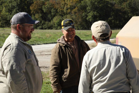 Three farmers stand in a field talking about the benefits of Vermeer balers